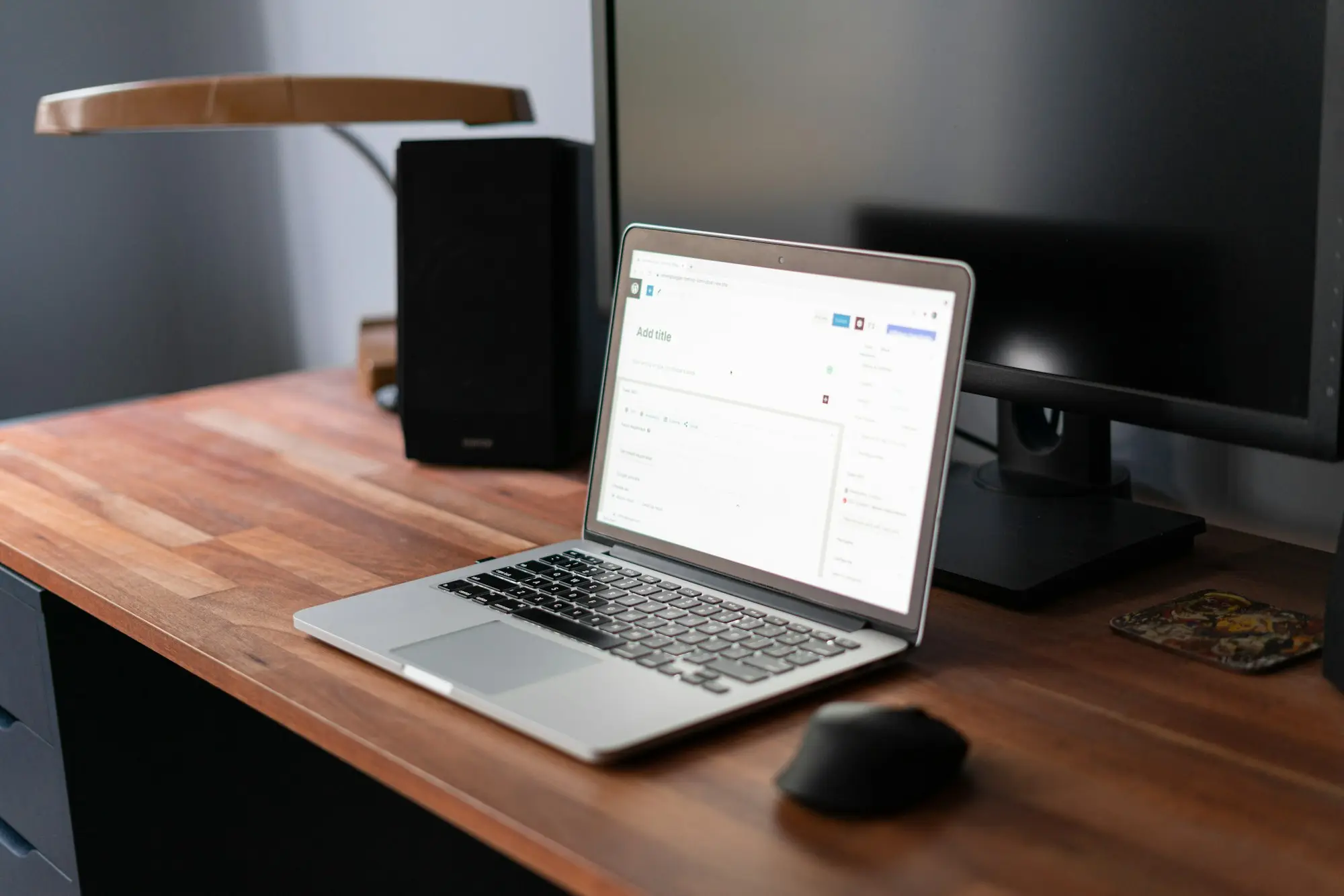 Un portatile acceso su una scrivania di legno, accanto a un monitor e a una lampada